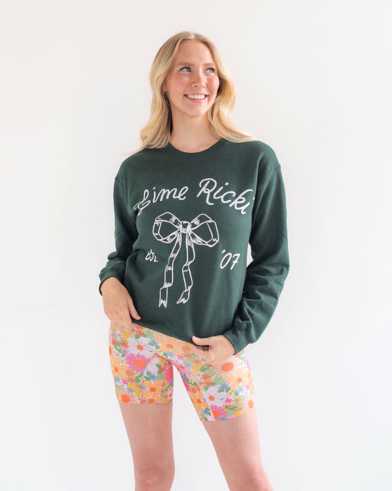 Photo of woman wearing a green Lime Ricki crew neck sweatshirt with multi colored floral long swim shorts