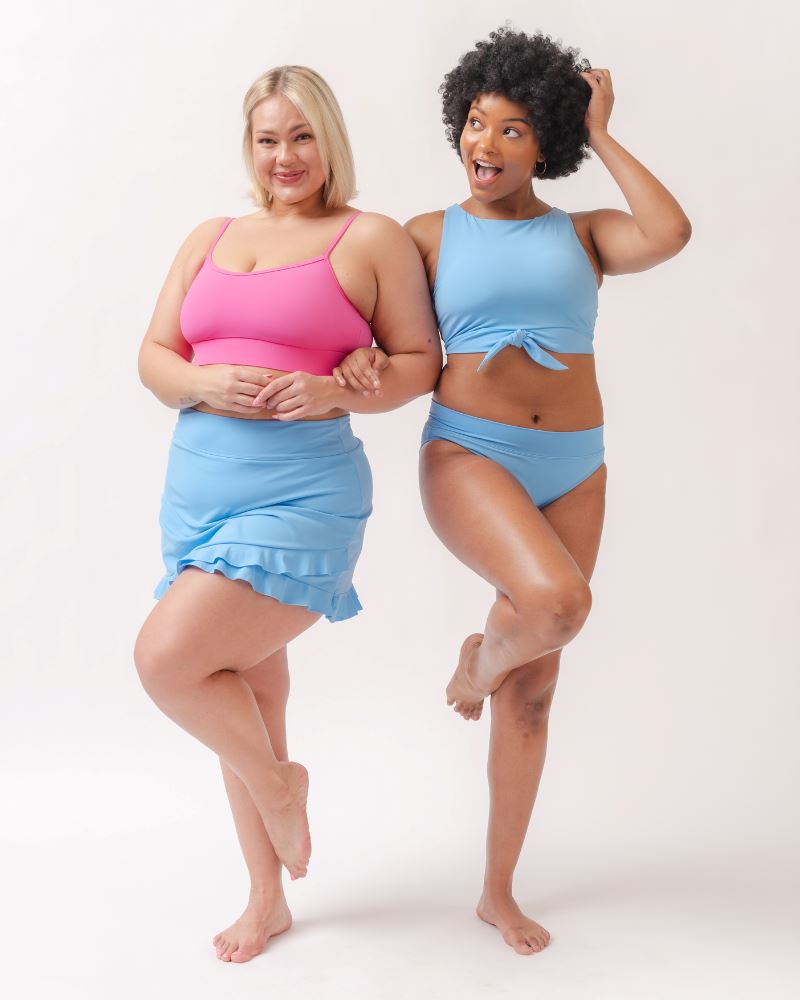 Photo of two women one wearing a pink cropped swim top with a light periwinkle swim skirt the other wearing a light periwinkle cropped swim top with a light periwinkle classic swim bottom