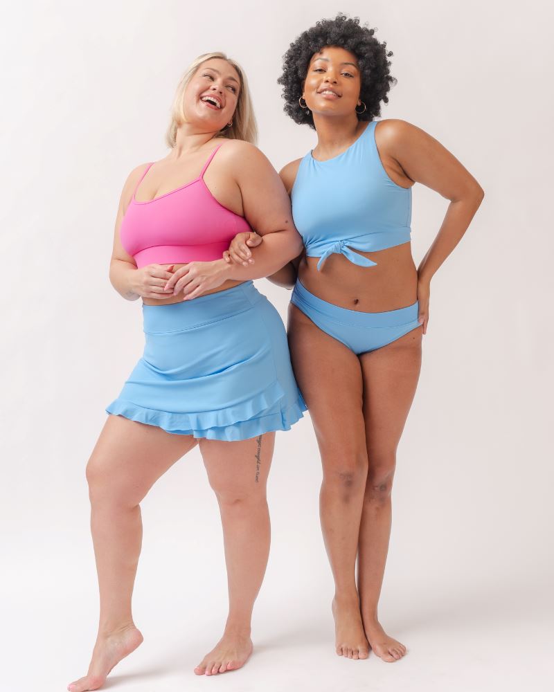 Photo of two women wearing a light periwinkle blue knotted swim crop top and a light periwinkle blue swim bottom and a dark pink swim bralette and a light periwinkle blue swim skirt
