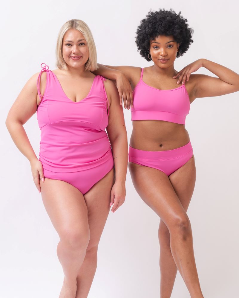 Photo of two women one with her hand on her hip wearing a pink swim tankini with pink low waist swim bottoms and the other wearing a pink cropped swim top with pink low waist swim bottoms