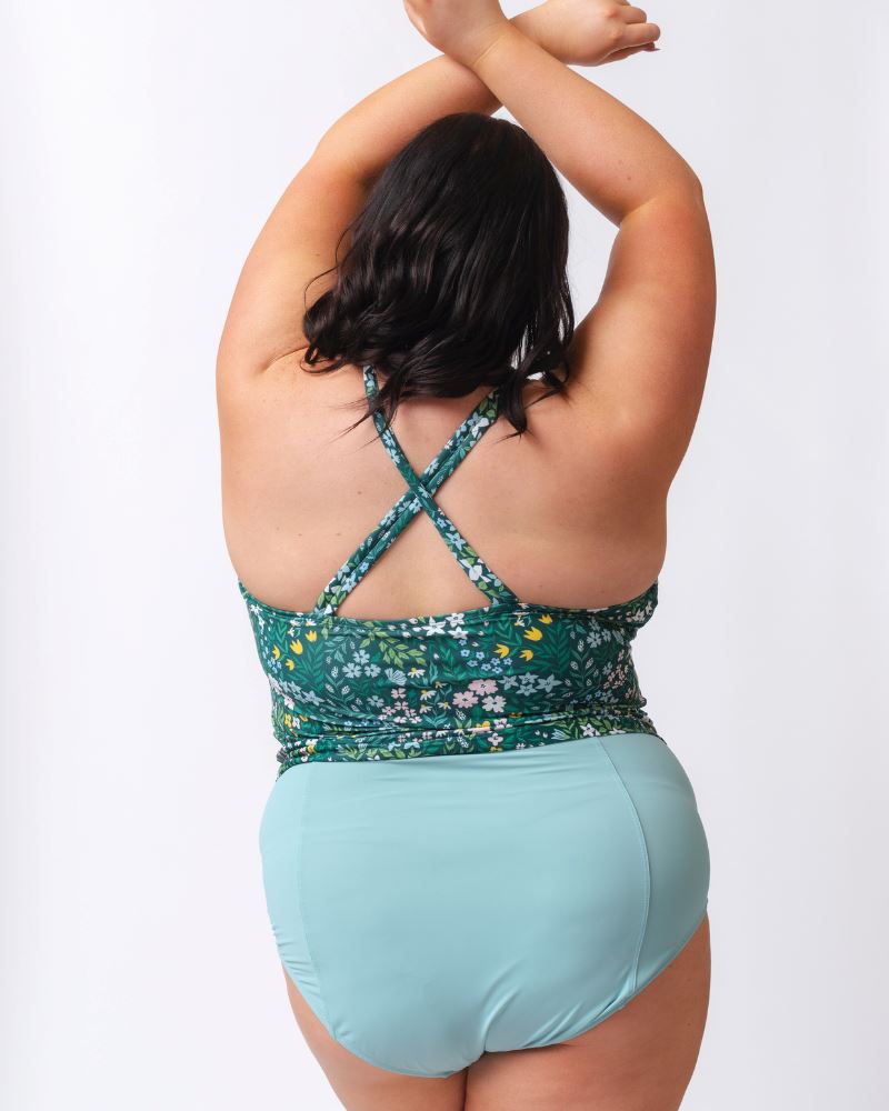 Photo of a woman wearing a dark green floral double-cinch tankini swim top and a light blue high waist swim bottom- back angle