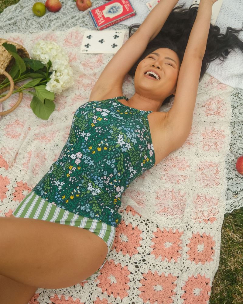Photo of a woman laying down wearing a dark green floral double-cinch tankini swim top and a green and white striped/ pink and white floral reversible swim bottom- striped side