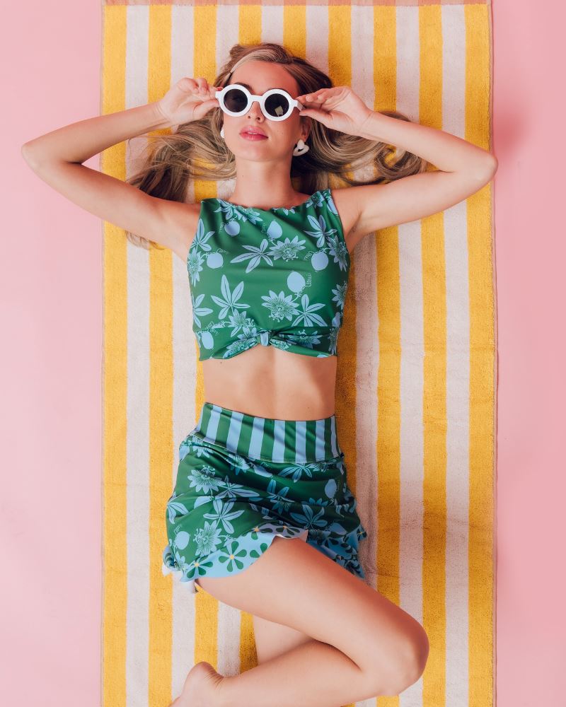 Photo of a woman laying on a towel wearing a green and blue floral swim crop top and a green and blue floral swim skirt bottom