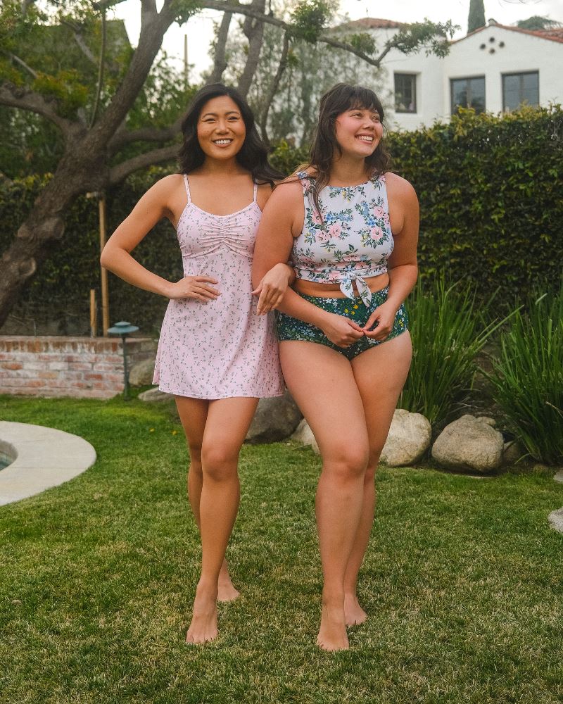 Photo of two women posing together wearing a pink floral swim dress and the other woman wearing a white and pink floral swim crop top and a dark green floral/ light green reversible swim bottom- floral side