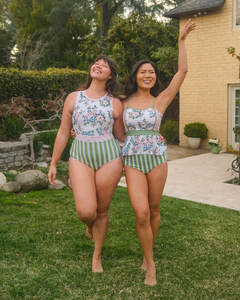 Photo of two women standing together one wearing a pink and white floral one piece swimsuit the other wearing a pink and white floral swim top with green and white stripe high waist swim bottoms