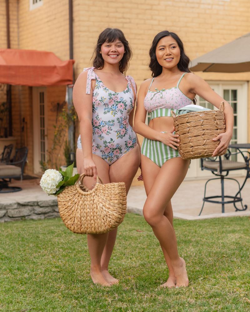Photo of two women standing together one wearing a pink and white floral one piece swimsuit the other wearing a pink floral cropped swim top with green and white stripe high waist swim bottoms
