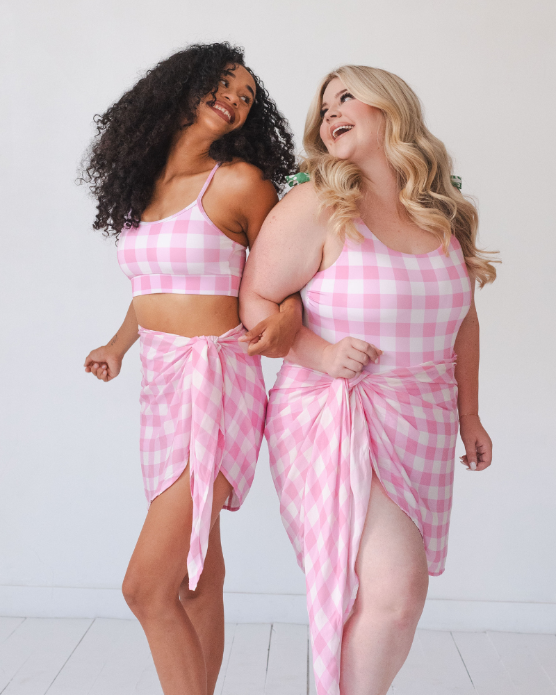Photo of two women linking arms smiling at each other one wearing a pink and white gingham cropped swim top with pink and white gingham sarong the other wearing a pink and white gingham one piece swim suit with pink and white gingham sarong