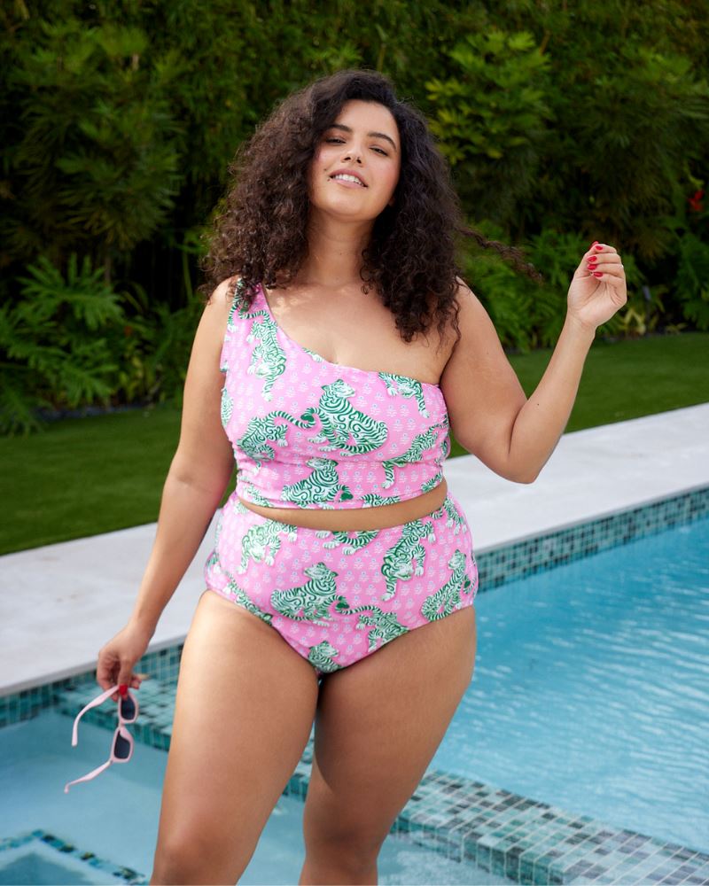 Photo of a woman standing by the pool wearing a bold pink and green tiger print one shoulder crop swim top with matching high waist swim bottoms