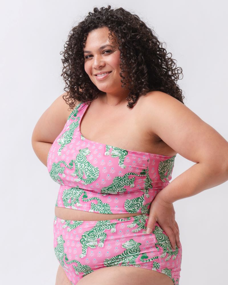 Photo of a woman with her hands on her hips wearing a bold pink and green tiger print one shoulder crop swim top with matching high waist swim bottoms
