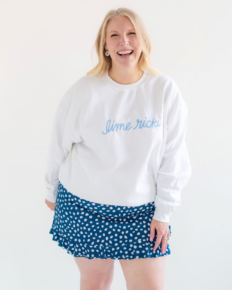 Photo of woman smiling wearing a Lime Ricki white crew neck sweatshirt with a blue and white polka dot swim skirt