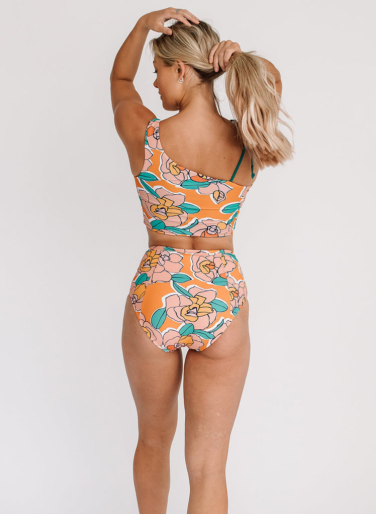 Photo of a woman with her back facing us and grabbing her hair while wearing an orange floral cropped swim top with orange floral high waist swim bottoms