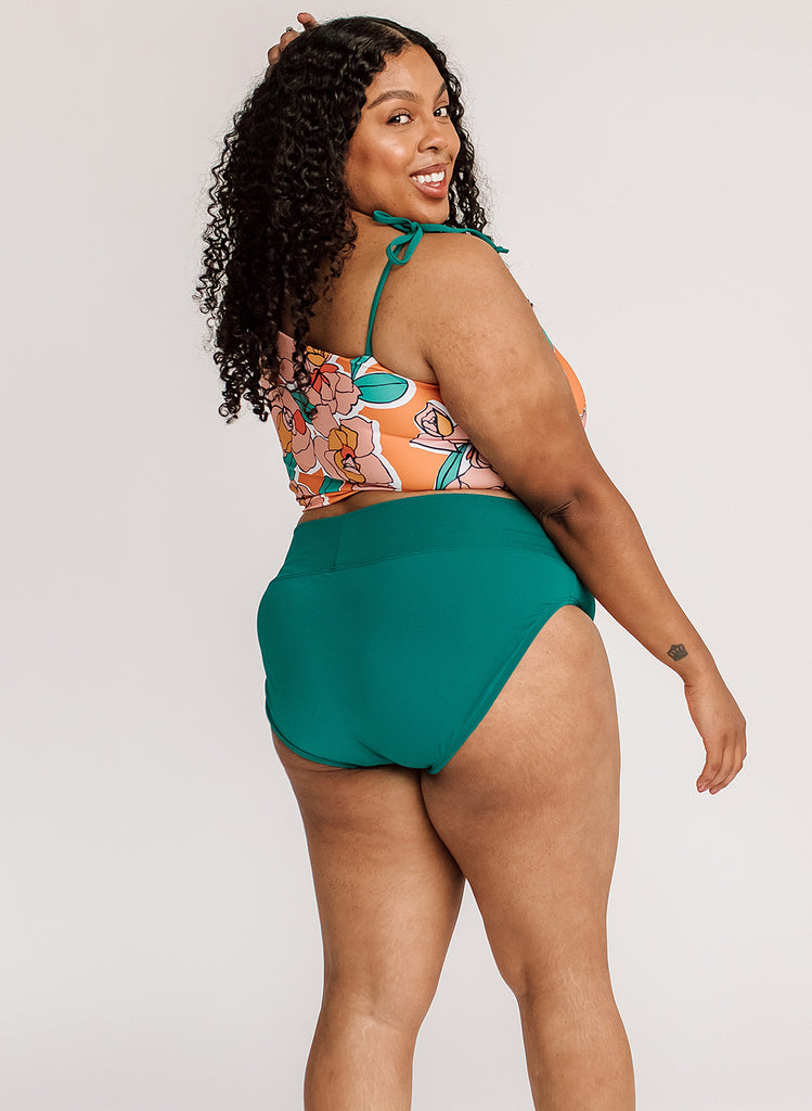 Photo of a woman with her back facing us looking over her shoulder while wearing an orange floral cropped swim top with blue classic low rise swim bottoms