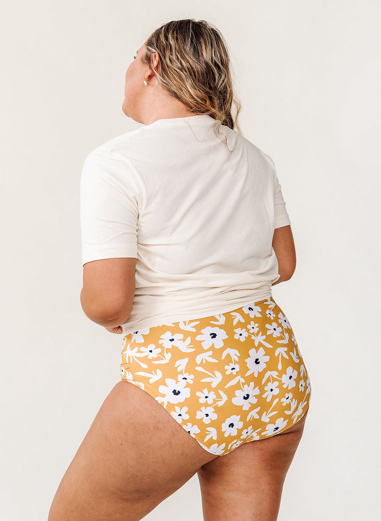 Photo of a woman wearing a white graphic tee shirt and a yellow floral swim bottom- back angle