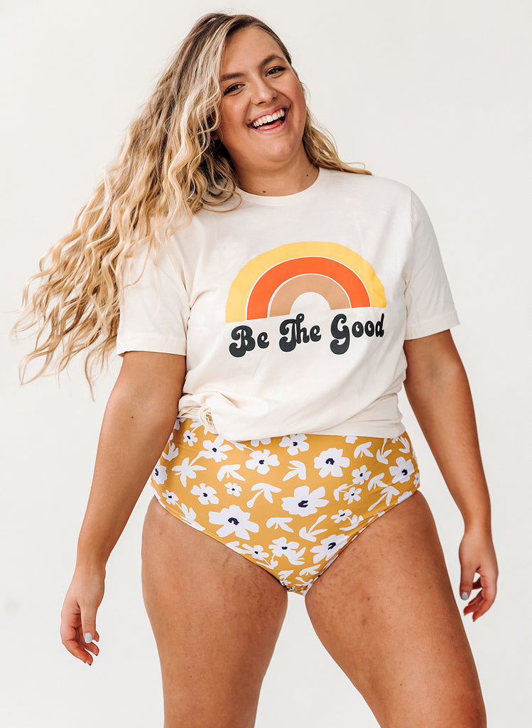 Photo of woman smiling while wearing a white "Be The Good" t-shirt with yellow floral high waist swim bottoms