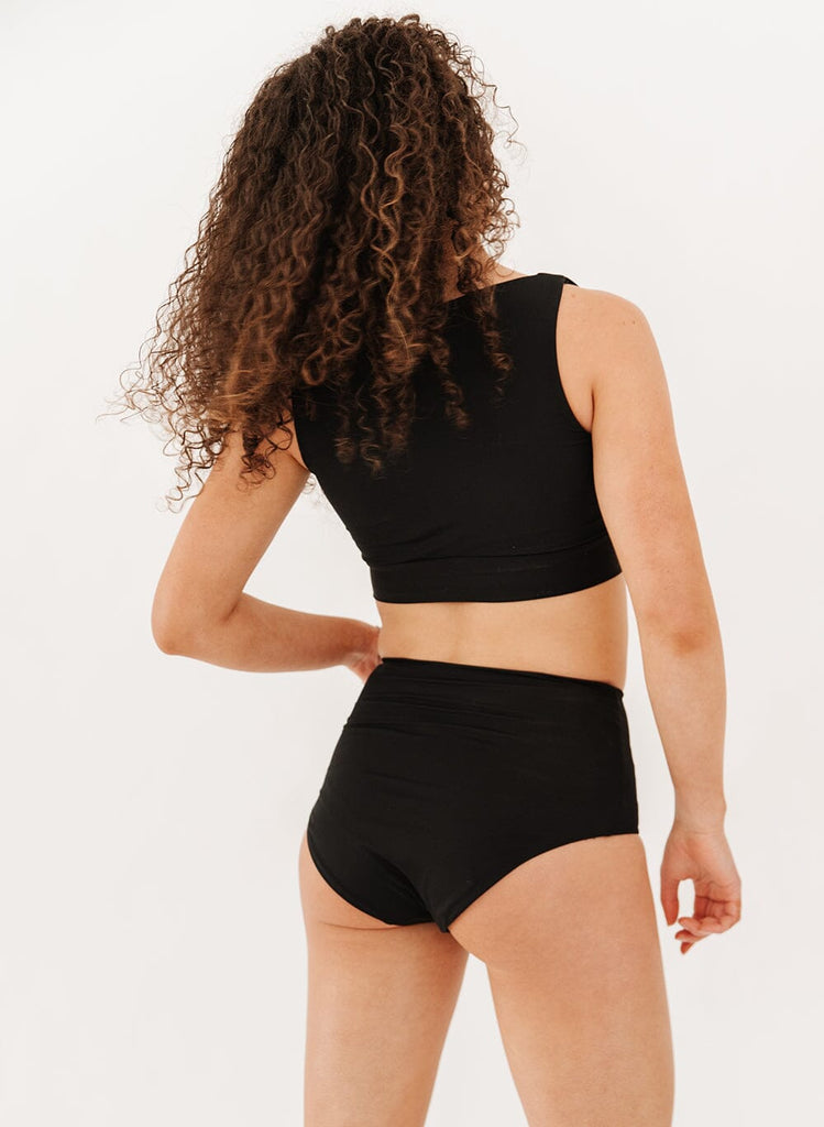 Photo of a woman wearing black ultra high-waist swim bottoms with a black knotted crop swim top back angle