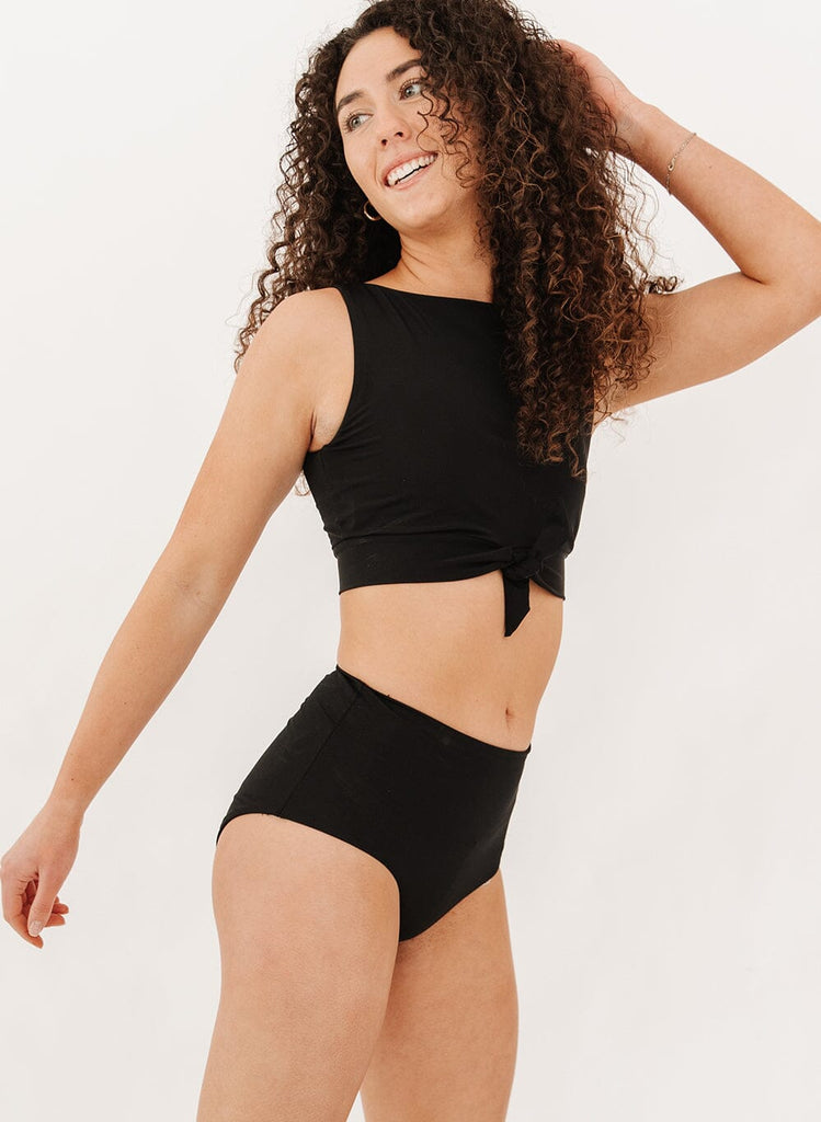 Photo of a woman wearing black ultra high-waist swim bottoms with a black knotted crop swim top side angle