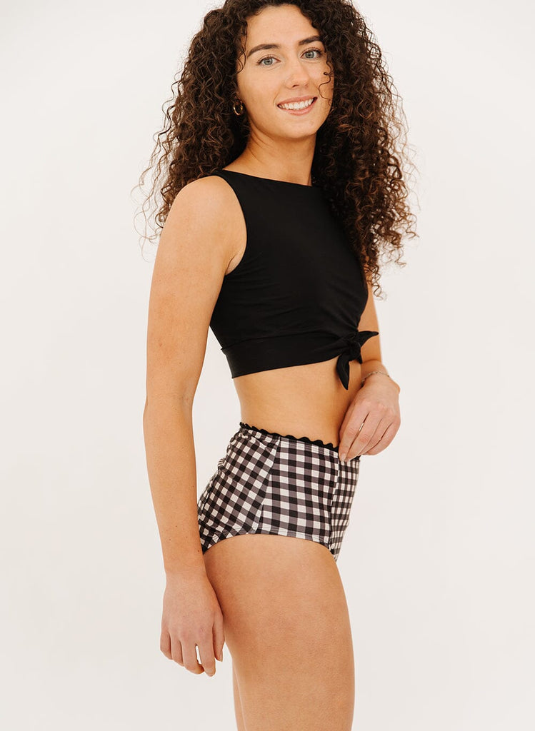 Photo of a woman wearing black gingham high-waist swim bottoms and a black knotted swim crop top side angle