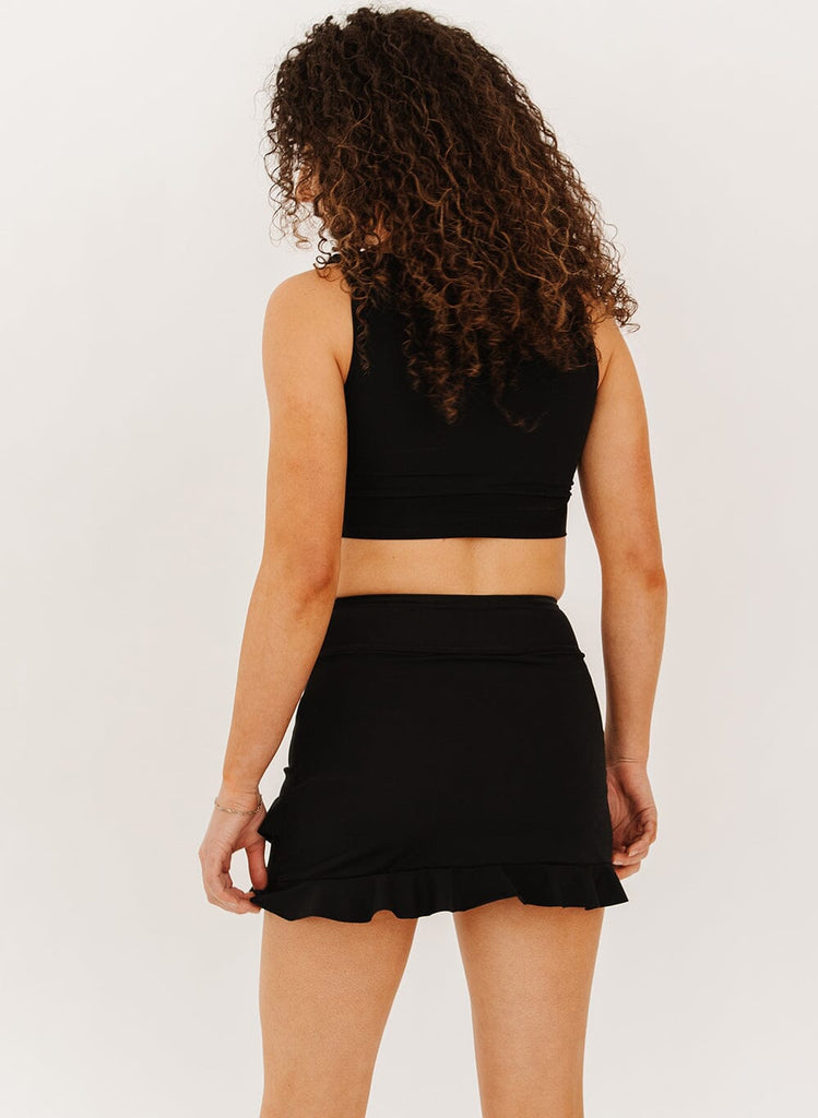 Photo of a woman wearing a black high-waist ruffle swim skirt with a black knotted crop swim top back angle