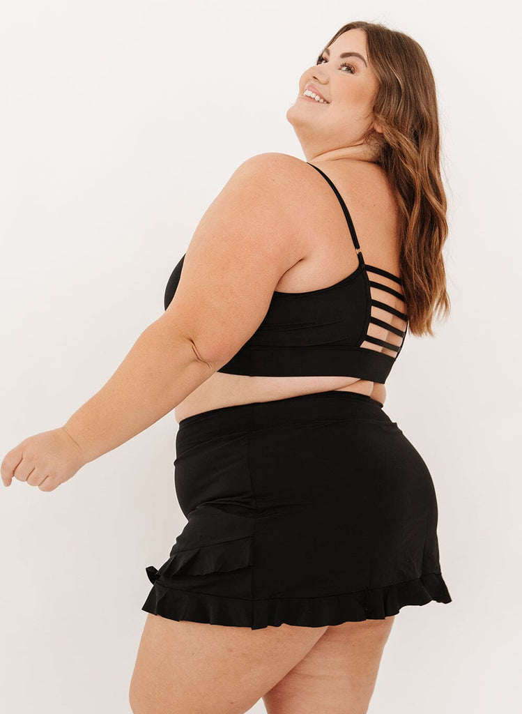 Photo of a woman wearing a black high-waist ruffle swim skirt with a black swim bralette side angle