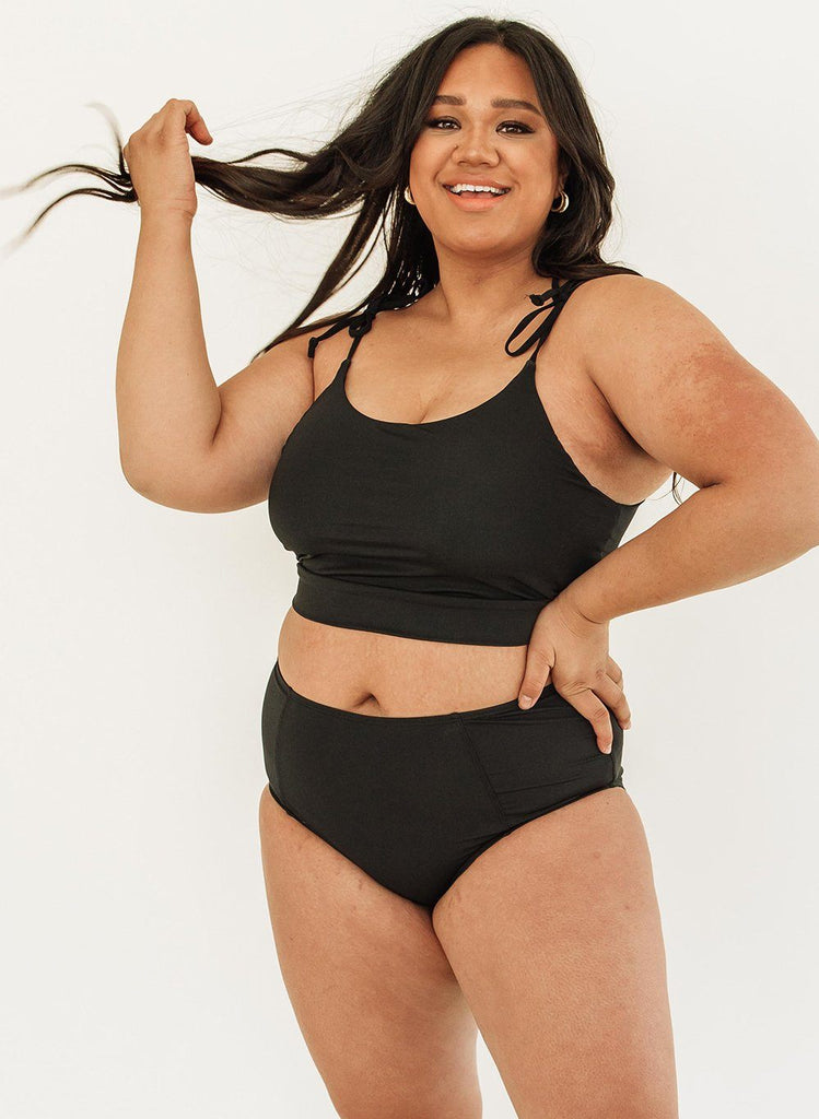Photo of woman posing with hand on hip while wearing a black cropped swim top with black high waist swim bottoms