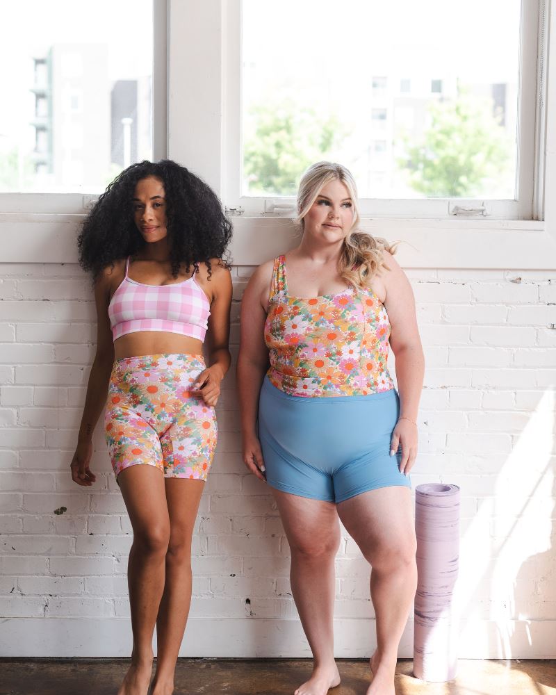 Photo of two women one wearing a pink gingham bralette swim top with multi colored long swim shorts and the other wearing a multi colored square neck swim top with blue swim shorts
