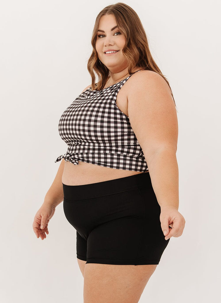 Photo of a woman wearing high-waist black swim shorts and a black gingham knotted crop swim top side angle