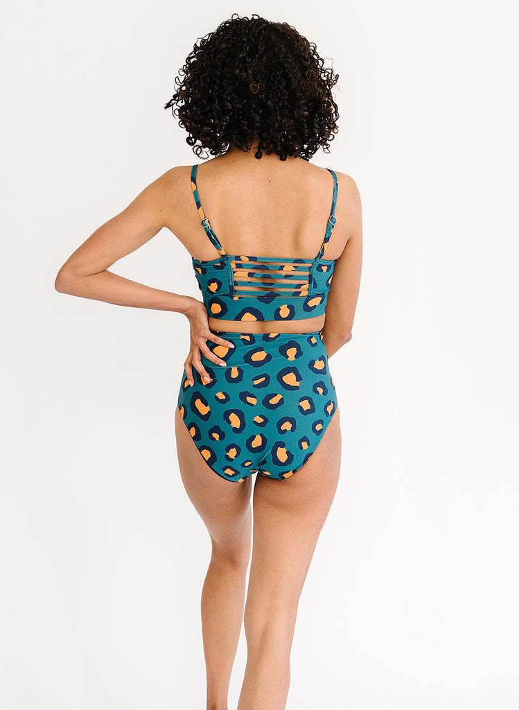 Photo of a woman with her back facing us and her hand on her hip while wearing a blue leopard print cropped swim top with blue leopard print high waist swim bottoms