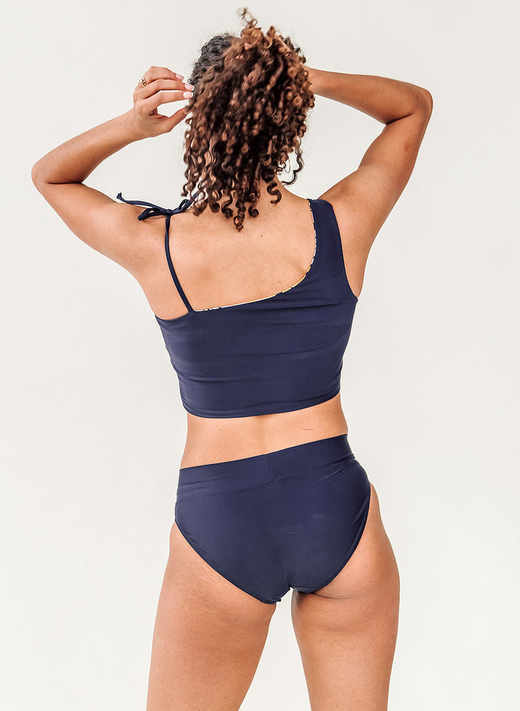 Photo of a woman with her back facing us and her hands in her hair while wearing a blue cropped swim top with blue classic low rise swim bottoms