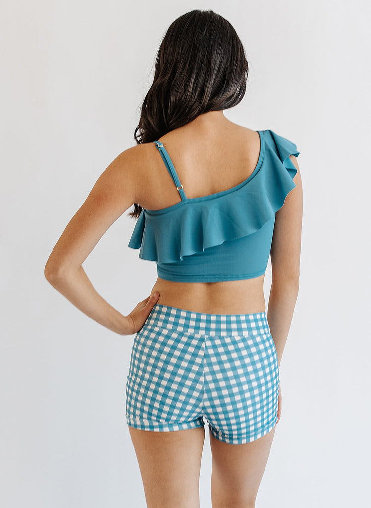 Photo of woman wearing blue ruffle cropped swim top with blue and white gingham swim shorts back angle