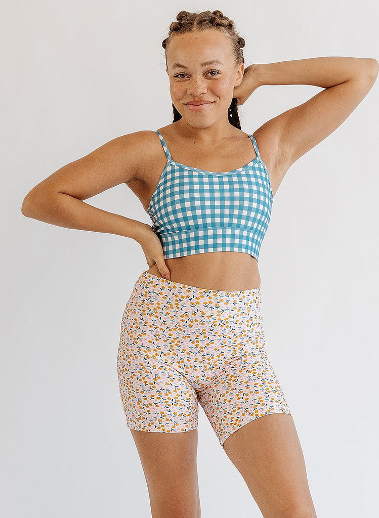 Photo of woman wearing blue and white gingham bralette swim top with multi color floral long swim shorts
