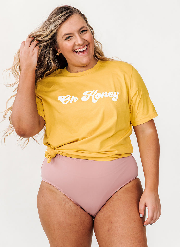 Photo of a woman wearing a yellow "Oh Honey" graphic tee shirt and an orchid pink swim bottom