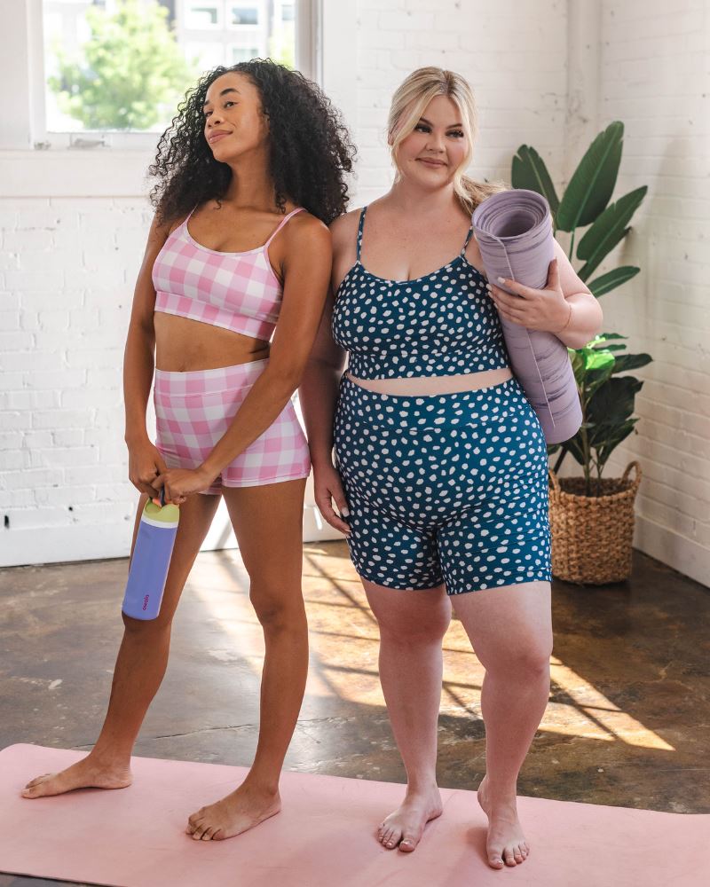 Photo of two women one wearing a pink gingham bralette swim top with pink gingham swim shorts the other wearing a blue and white dot bralette swim top with blue and white dot long swim shorts