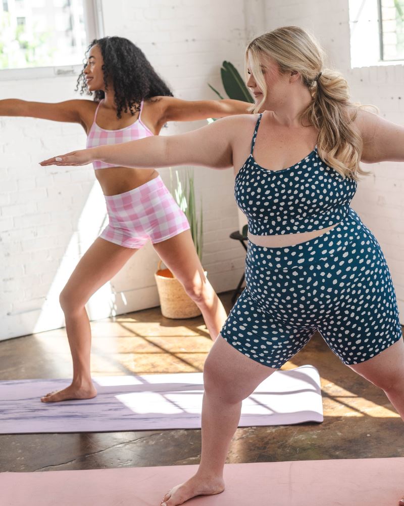Photo of two women doing yoga one wearing a pink gingham bralette swim top with pink gingham swim shorts the other wearing a blue and white dot bralette swim top with blue and white dot long swim shorts