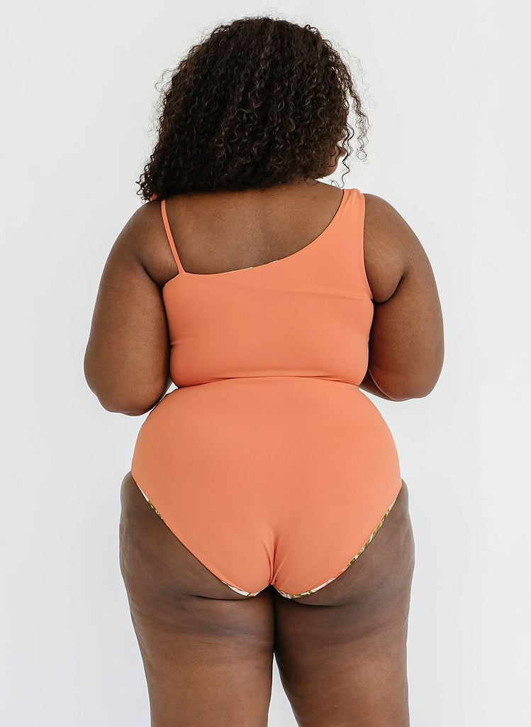 Photo of a woman wearing a sandstone orange one-shoulder swim crop top and a sandstone orange swim bottom- back angle