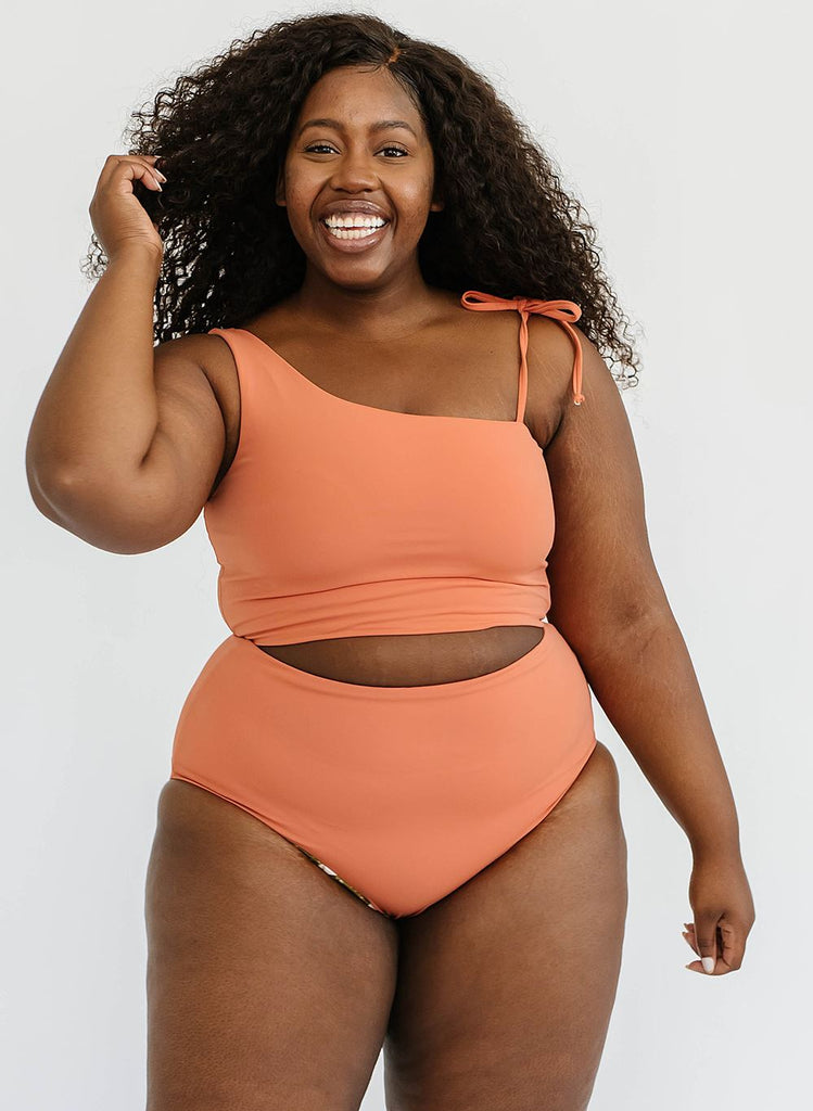 Photo of a woman wearing a sandstone orange one-shoulder swim crop top and a sandstone orange swim bottom