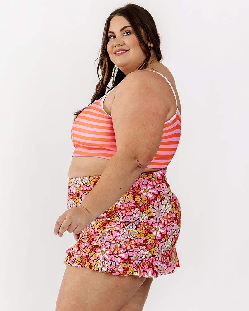 Photo of a woman wearing a groovy Blooms floral swim skirt bottom and an orange and pink stripe swim bralette side angle