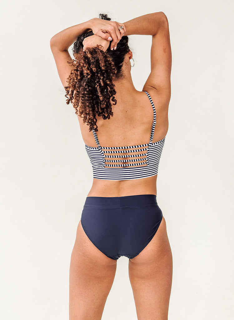 Photo of a woman with her back facing us wearing a blue and white striped cropped swim top with blue classic low rise swim bottoms