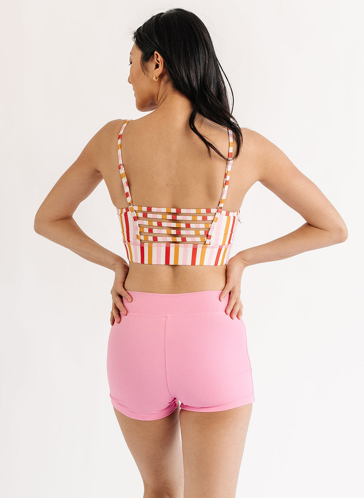 Photo of woman wearing multi color stripe bralette swim top with pink swim shorts back angle