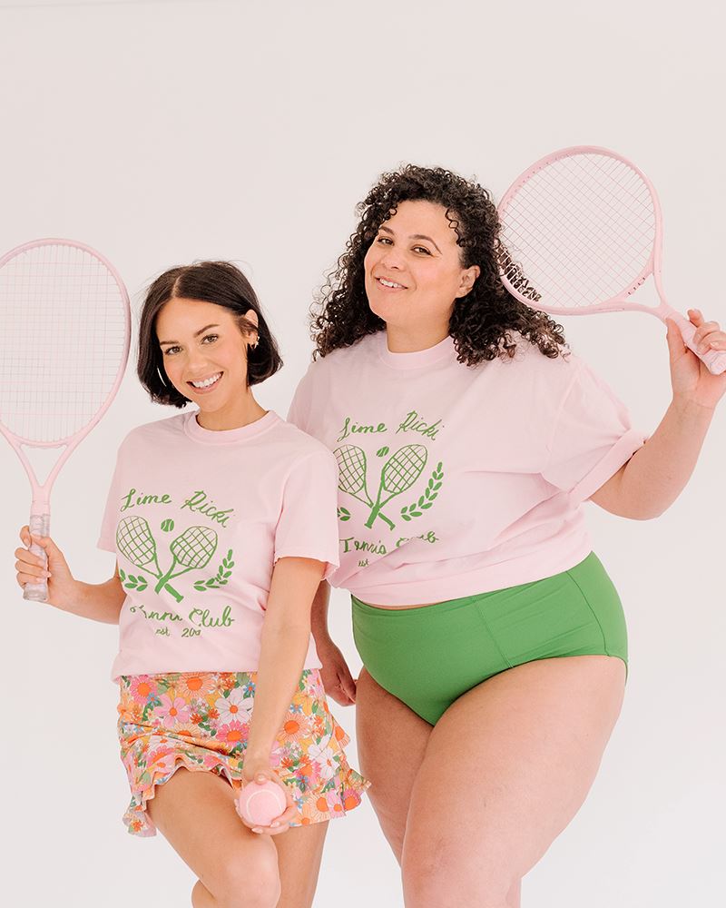 Photo of two women holding tennis rackets one wearing a lime ricki tennis club graphic tee with a multi colored floral swim skirt the other wearing a lime ricki tennis club graphic tee with green swim bottoms