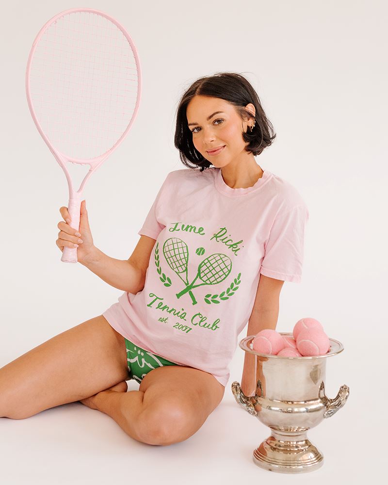 Photo of woman holding tennis racket wearing lime ricki tennis club graphic tee with green and white floral swim bottoms