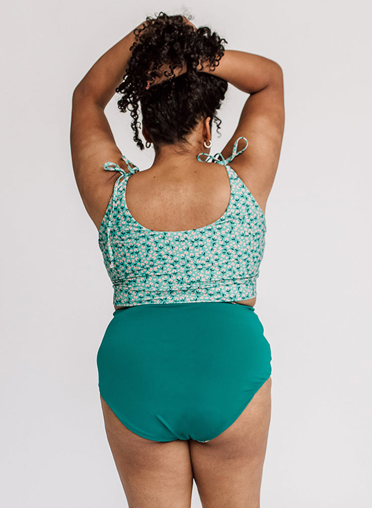 Photo of a woman with her back facing us and her hands in her hair while wearing a blue floral cropped swim top with blue high waist swim bottoms