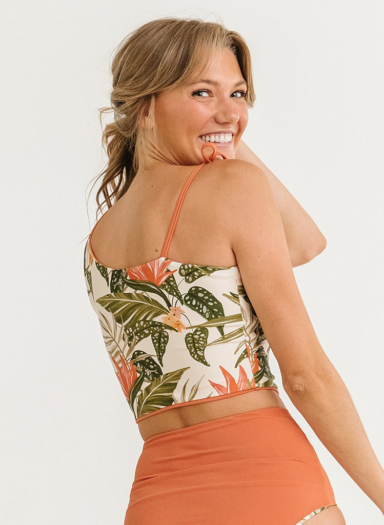 Close up photo of a woman with her back facing us smiling over her shoulder while wearing a green floral cropped swim top with orange high waist swim bottoms