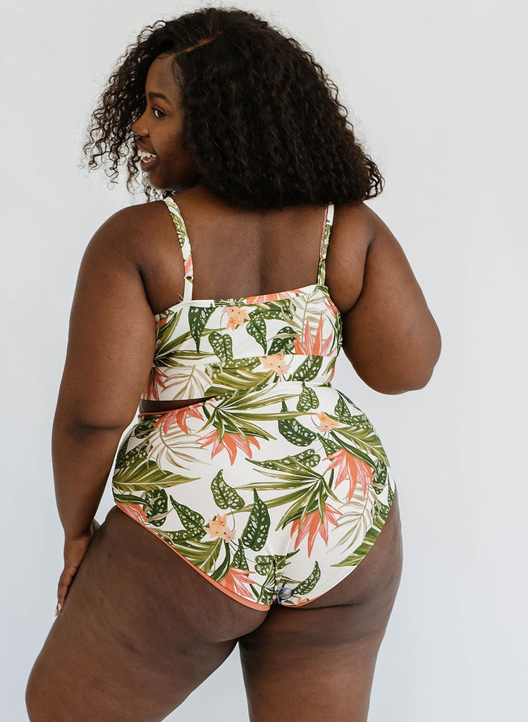 Photo of a woman with her back facing us and looking over her shoulder while wearing a green floral cropped swim top with green floral high waist swim bottoms