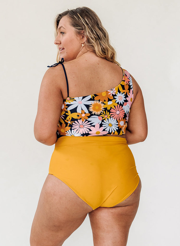 Photo of a woman wearing a multi colored floral one-shoulder swim crop top and yellow swim bottoms- back angle