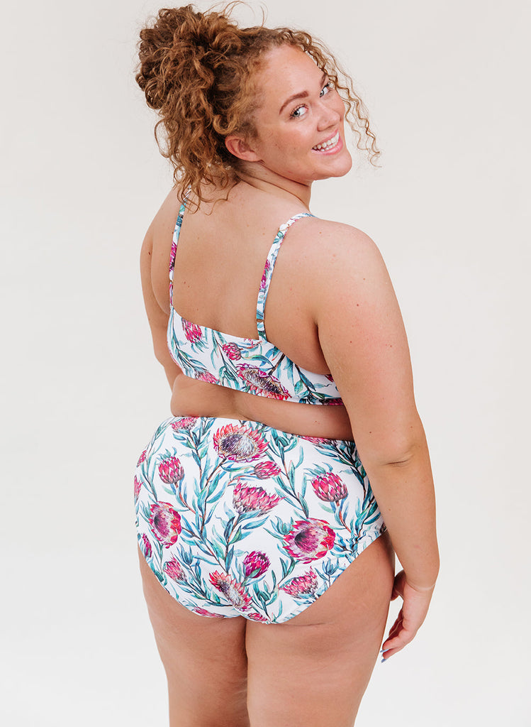 Photo of a woman with her back facing us looking over her shoulder while wearing a blue and pink floral cropped swim top with blue and pink floral high waist swim bottoms